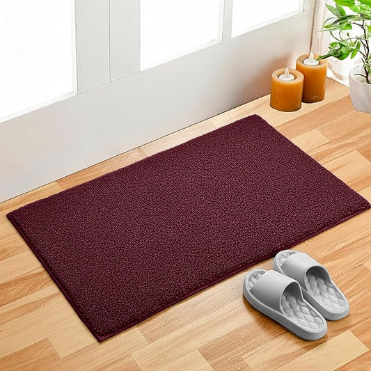 Maroon bathmat on wooden floor near door, with gray slippers, candles, and potted plant