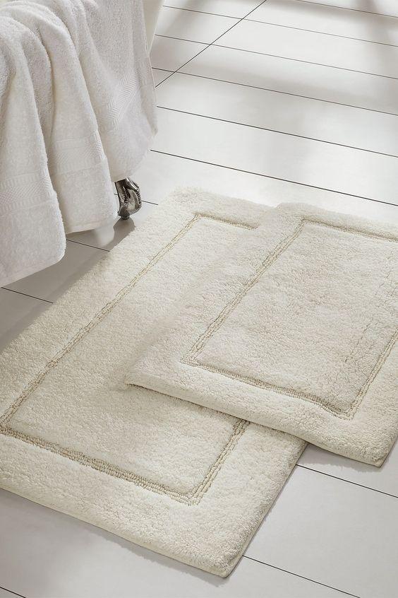 Plush cream bathmats on white tile floor next to a neatly draped bedspread