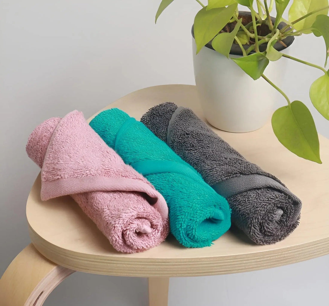 Rolled face towels in pink, teal, and gray on a wooden table beside a potted plant