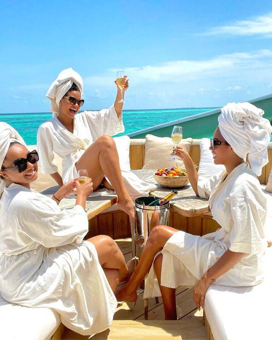 Three women in white bathrobes and towels relax on an outdoor patio, enjoying drinks by the ocean.