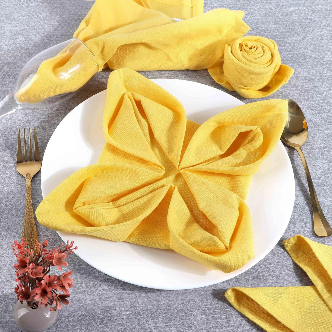 Yellow cotton napkin folded flower-shaped on white plate, gold cutlery, elegant table setting