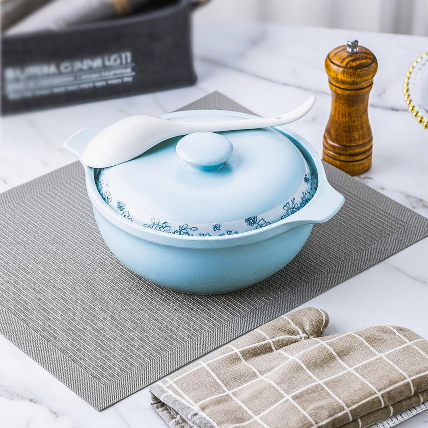 Light blue ceramic casserole dish with floral pattern, white spoon, and oven mitt on kitchen table