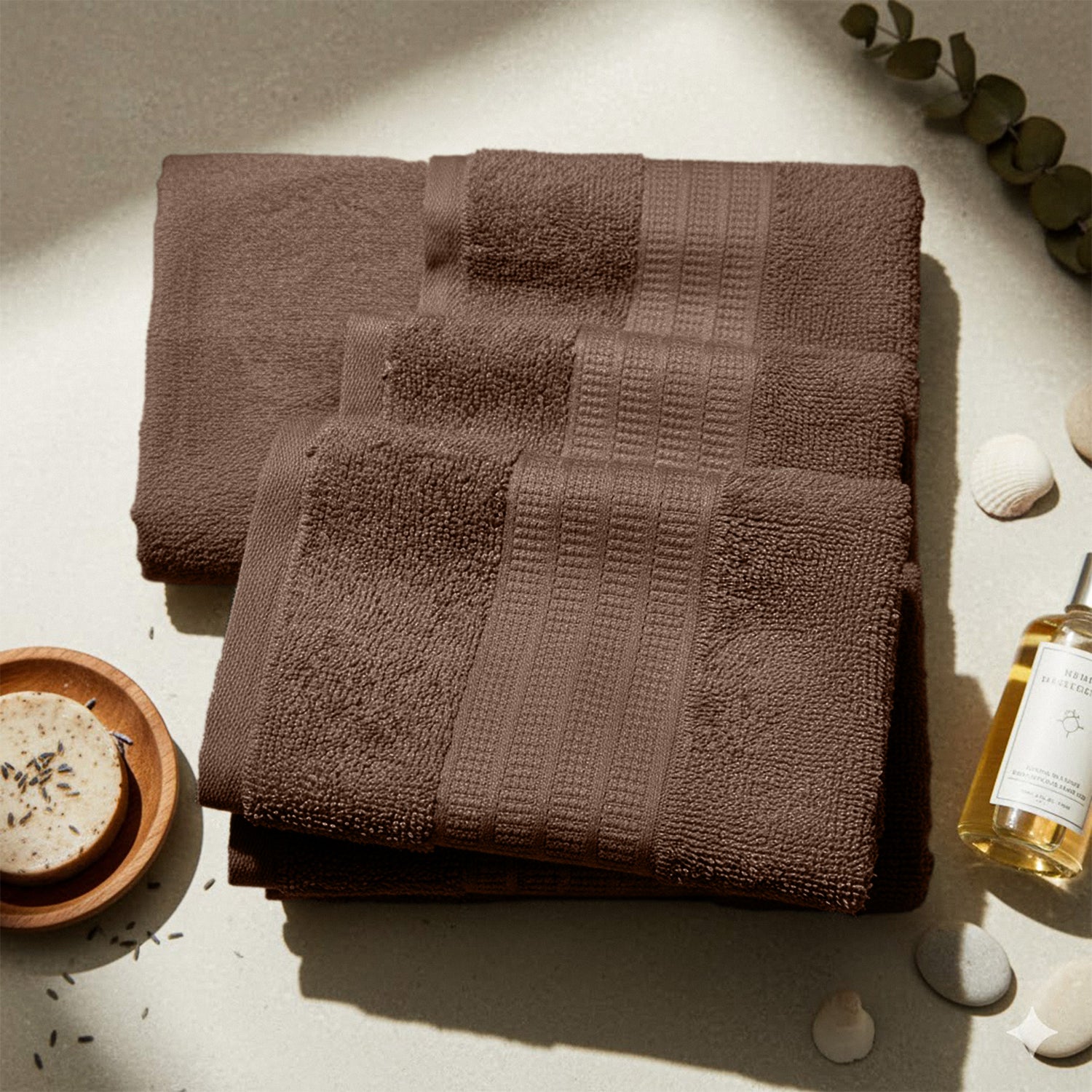 Set of brown bath towels on a light surface with soap, bottle, and decorative shells nearby