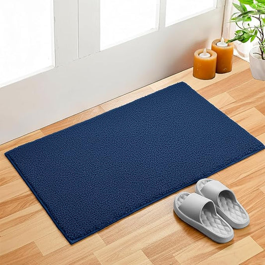 Navy blue bathmat on wood floor by door, with candles, plant, and gray slippers