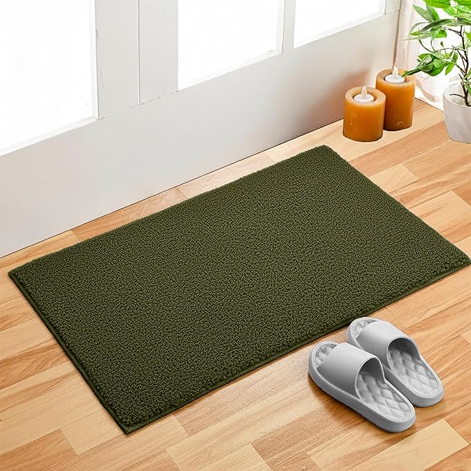 Green bathmat on wooden floor near door, with candles, plant, and gray slippers