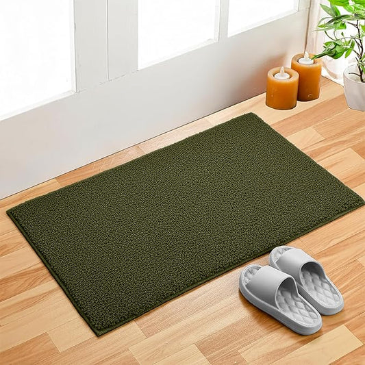Green bathmat on wooden floor near door, with candles, plant, and gray slippers
