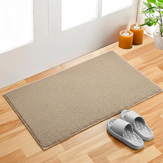 Beige bathmat on wooden floor near door, with gray slippers and candles beside a potted plant.