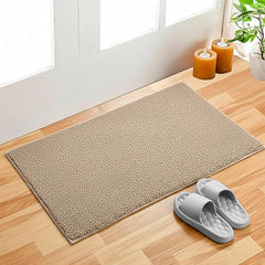 Beige bathmat on wooden floor near door, with gray slippers and candles beside a potted plant.