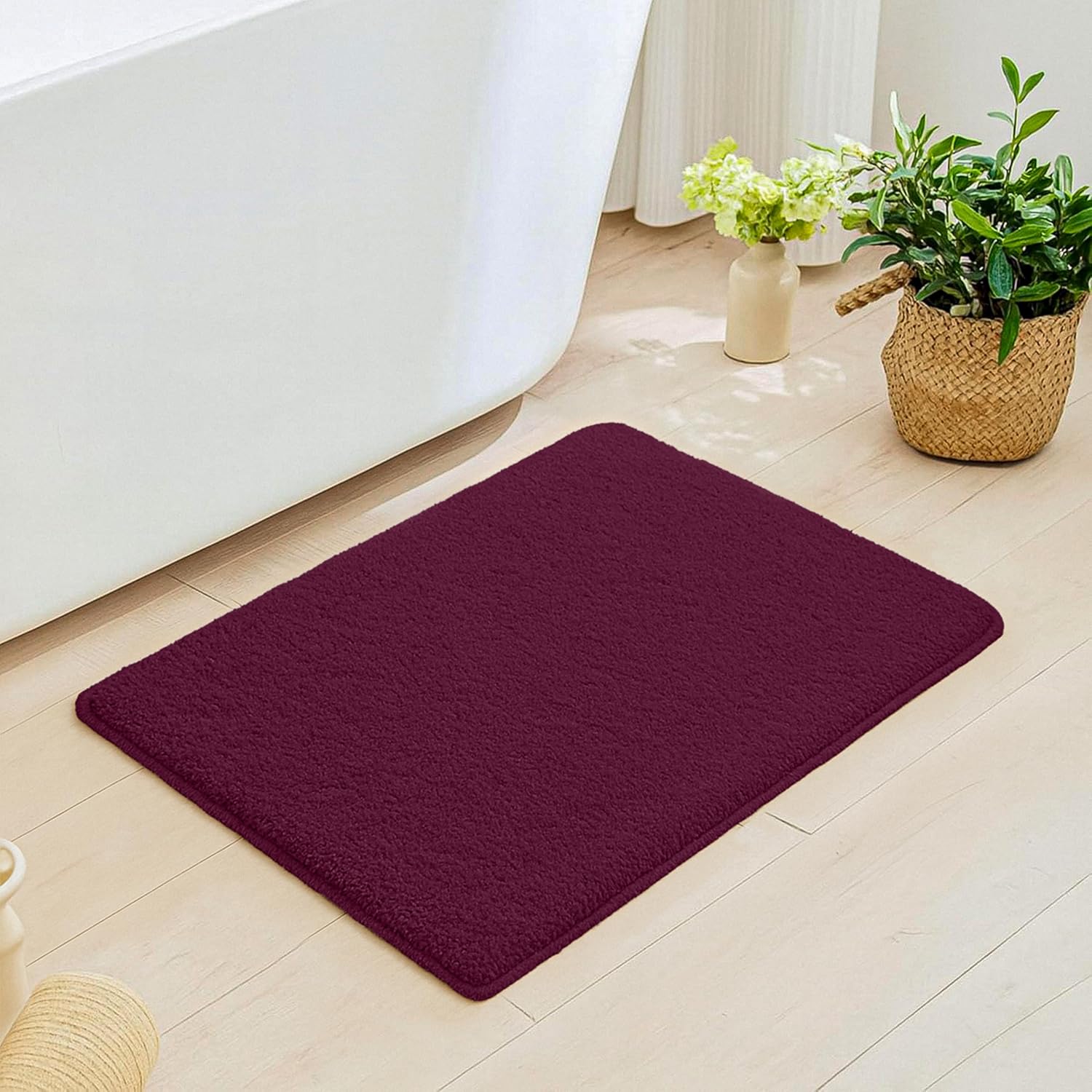 Plush maroon bathmat on light wood floor next to white bathtub with plants nearby