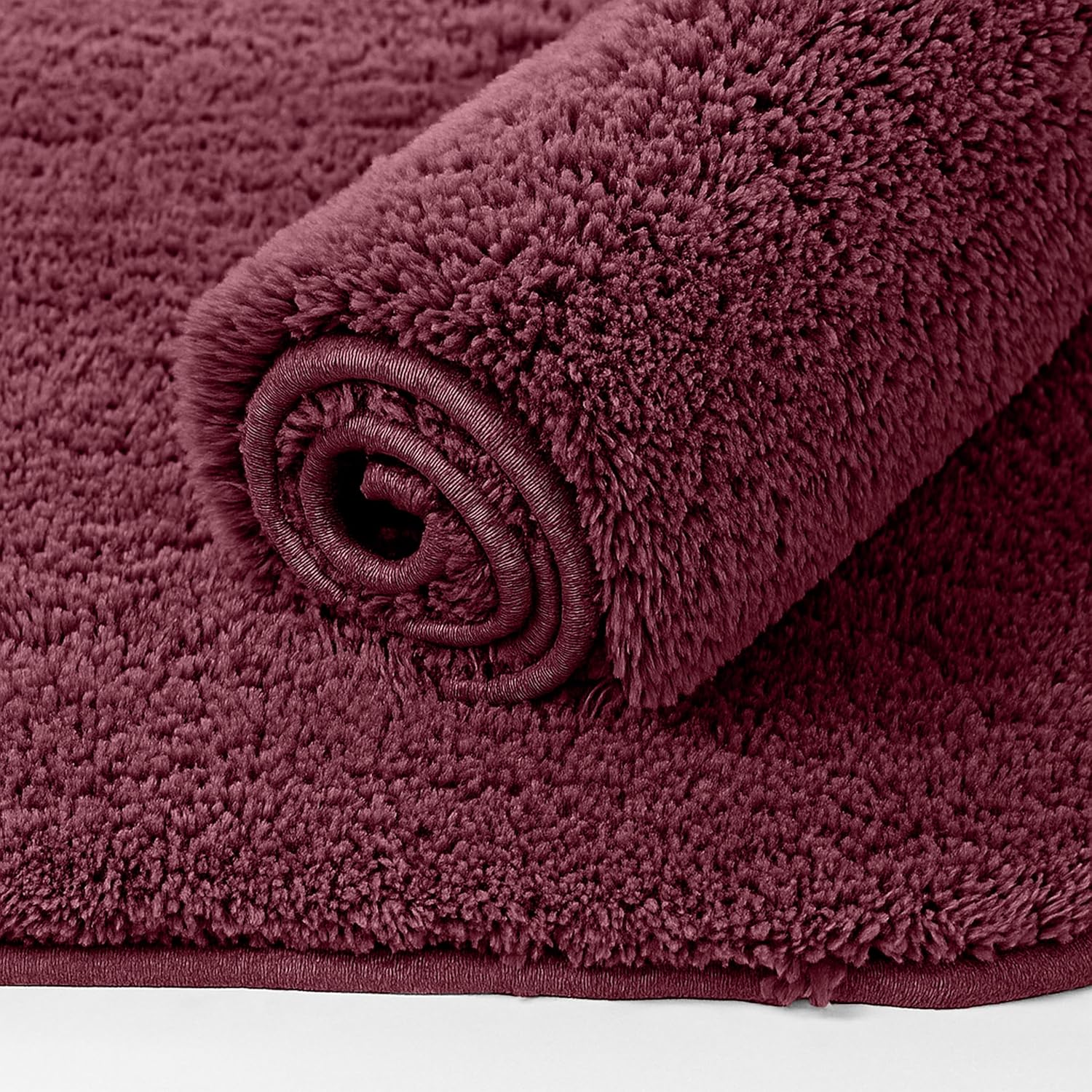 Soft maroon bathmat with plush textured surface, partially rolled on a white background